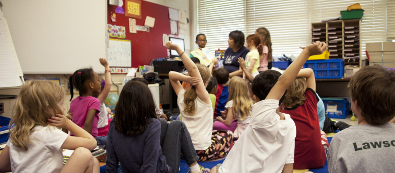 Group of gradeschoolers in classroom gathered around teacher for story time